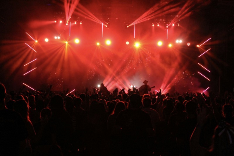 A crowd with hands raised facing a stage bathed in red light, a guitarist in a cowboy hat at the centre.