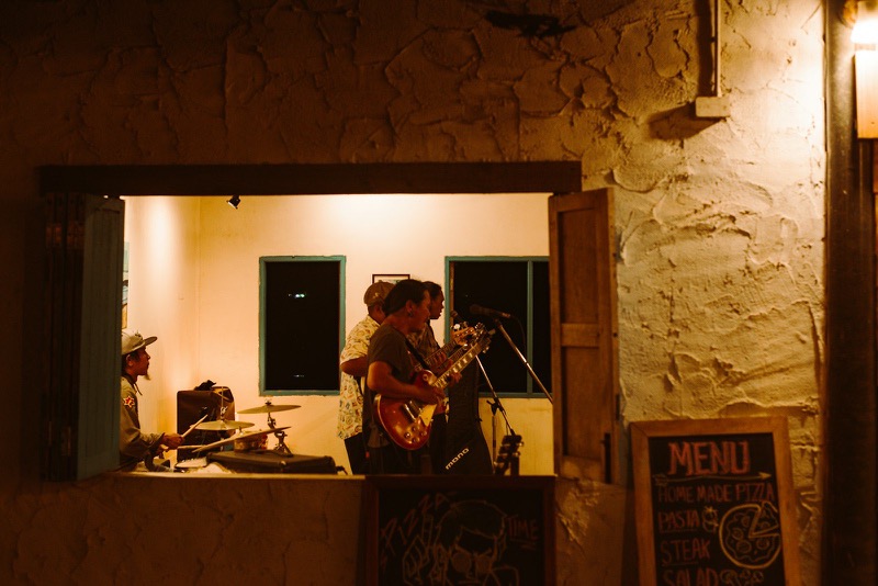 A band playing inside a warmly lit room, seen at night through an open window.