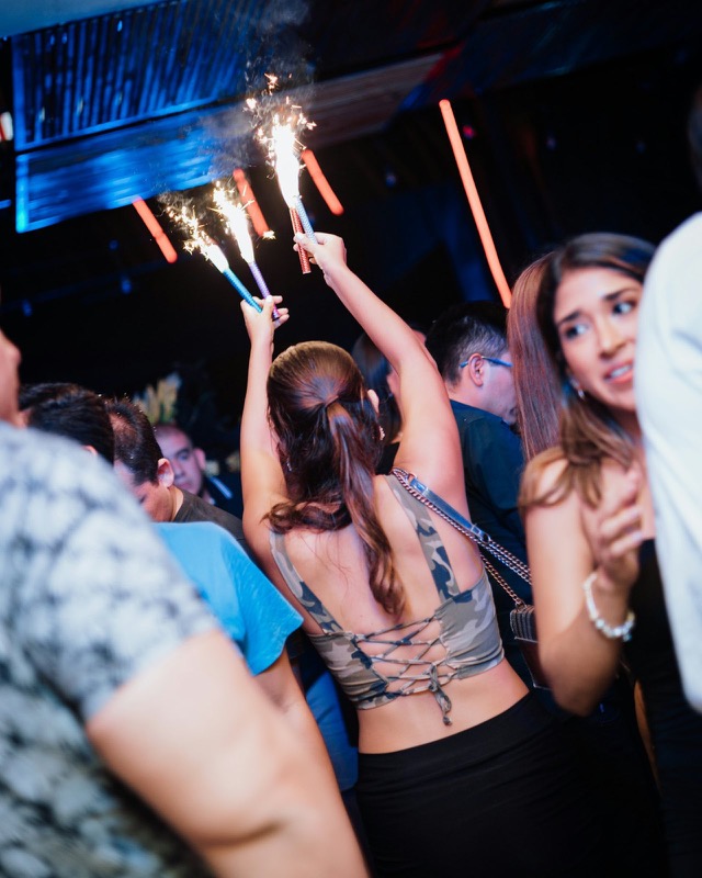 Woman holding three sparklers overhead at a club table, seen from behind.