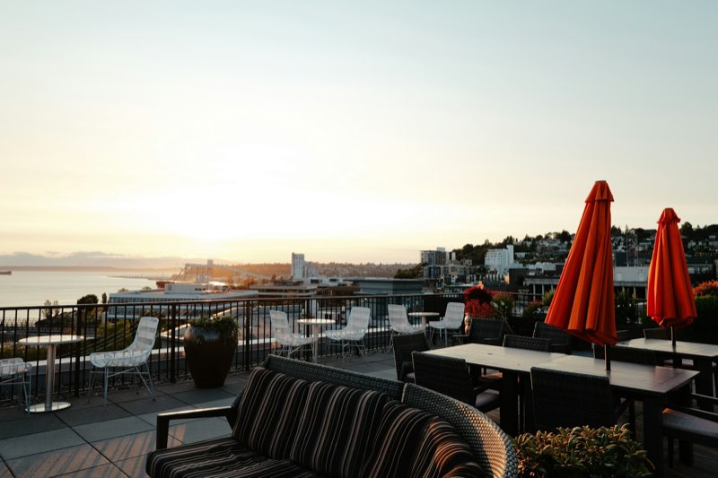 A rooftop patio with lounge seating and closed umbrellas overlooking water at sunset.