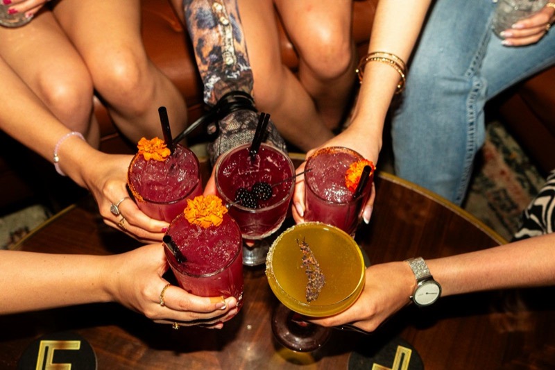 Five hands clinking cocktails with marigold garnishes over a round table.